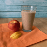 A peach smoothie in a clear glass with a teal background and peach colored cloth below next to peaches.