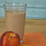 A dairy free peach smoothie in a clear cup with a full peach and peach slices next to it.