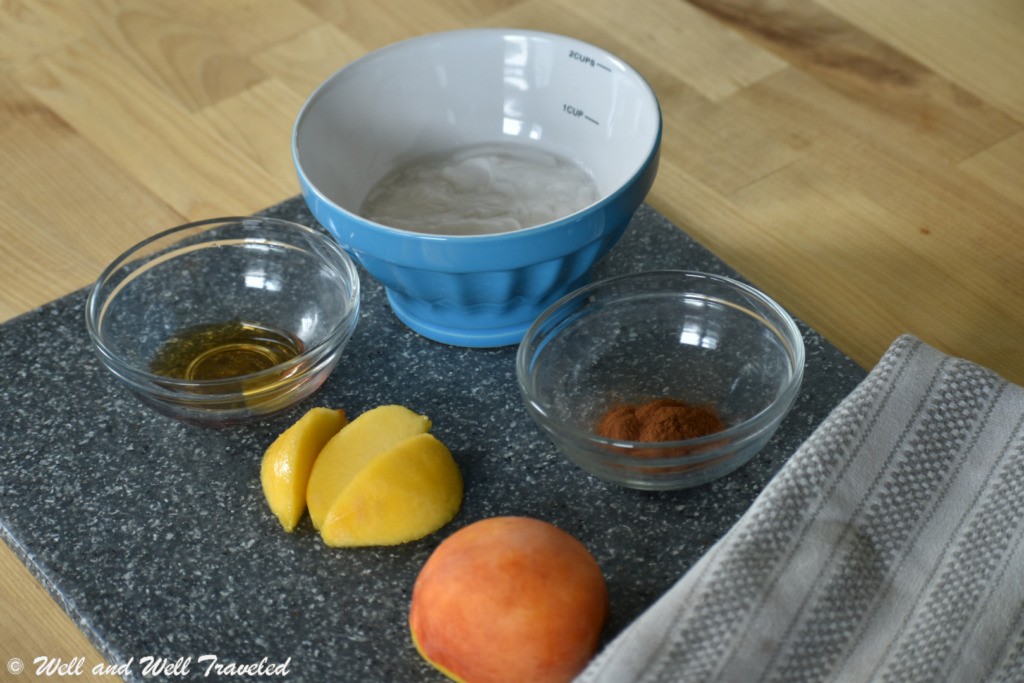 A peach and peach slices on a cutting board with cinnamon, maple syrup, and coconut cream in bowls next to them.. the ingredients for the dairy free peach smoothie