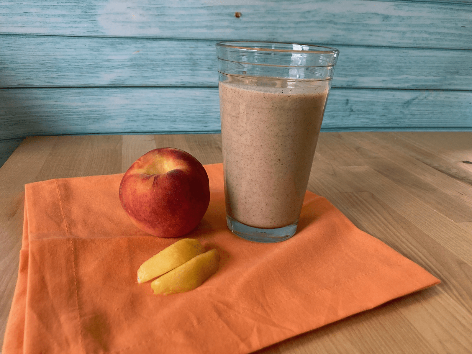 A peach smoothie in a clear glass with a teal background and peach colored cloth below next to peaches.