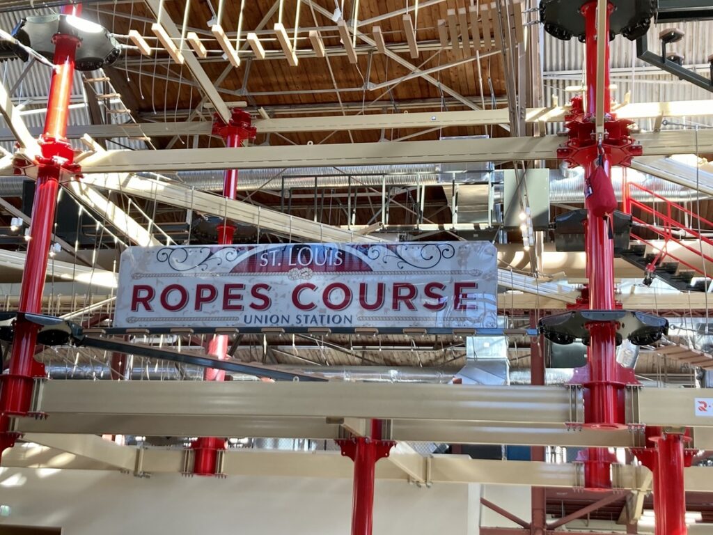 The inside of a large building. A sign saying "Ropes Course" is in front of a red ropes course, one of many family-friendly attractions in St. Louis