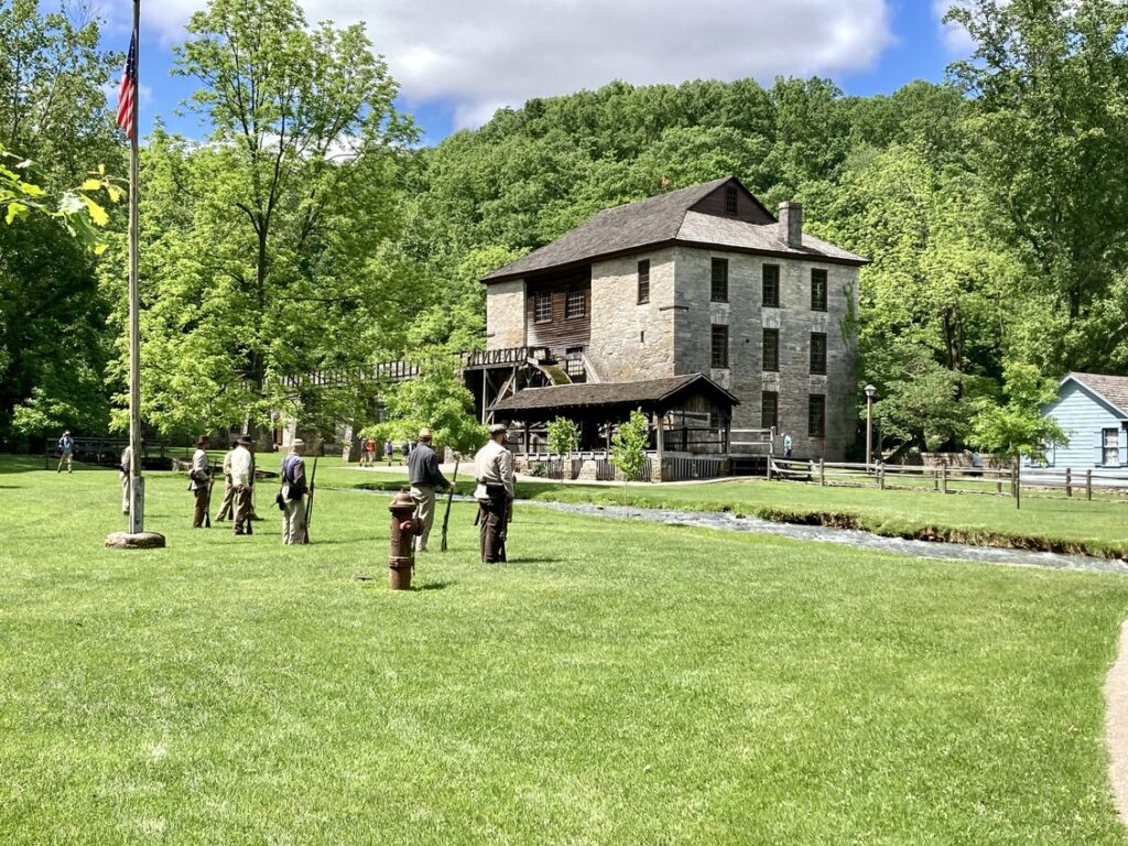 A pioneer village with a 3-story building with a mill attached in the background and men reenacting a war in the foreground