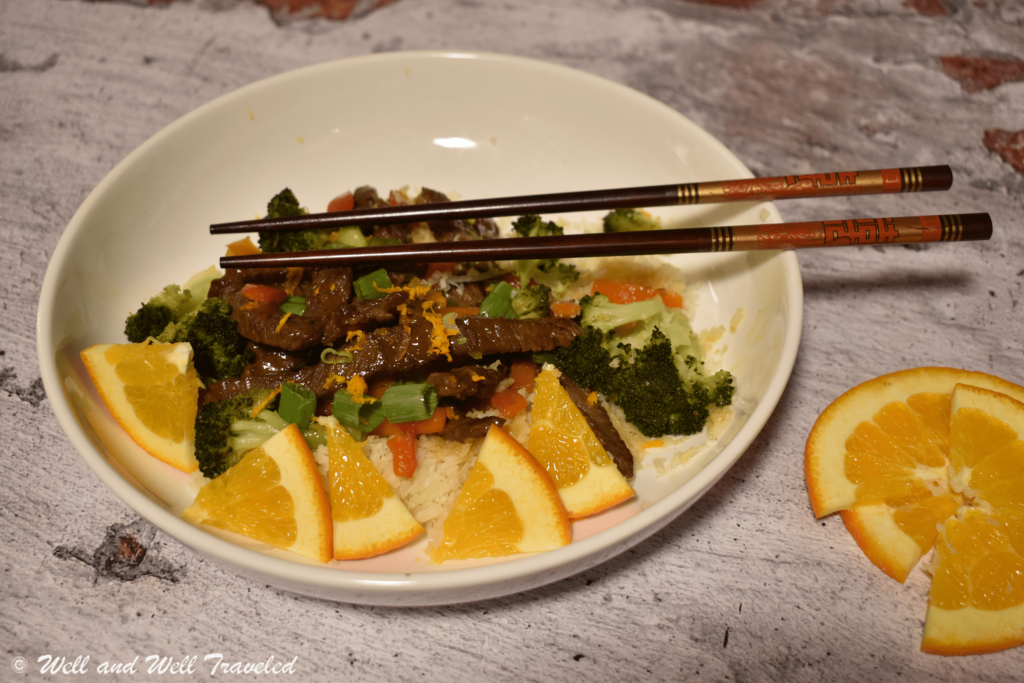 A white bowl with orange beef on cauliflower rice in it. Orange slices and chop sticks are decoratively placed